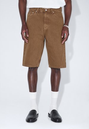 Mężczyzna ubrany w brązowe jeansowe szorty do kolan, białe skarpetki, czarne wsuwane buty oraz srebrną bransoletkę na prawej ręce.