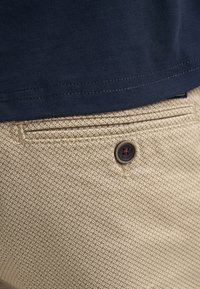 Gros plan d'une poche de pantalon beige texturé avec un bouton noir et un ourlet de chemise bleu marine au-dessus.