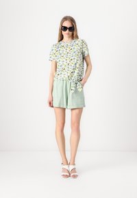 Blomstermønstret kortermet skjorte i hvit og grønn, kombinert med lysegrønne shorts med knytning i livet. Modell iført svarte solbriller og hvite sandaler.