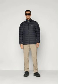 Μαύρο μπουφάν τύπου puffer με οριζόντιες ραφές, κλείσιμο με φερμουάρ και όρθιο λαιμό. Συνδυάζεται με μπεζ cargo παντελόνι και μαύρα παπούτσια.