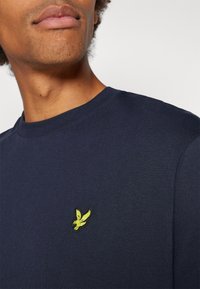 Granatowa bawełniana koszulka z okrągłym dekoltem i krótkimi rękawami, z żółtym wyszywanym logo ptaka na lewej piersi.