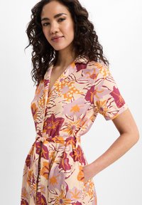 Abito floreale in crema con motivi rosa, arancioni e viola. Presenta maniche corte, colletto e una cintura in vita. Include tasche.
