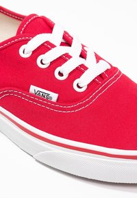 Zapatilla de lona roja con cordones blancos, ojales redondos y suela a rayas rojas y blancas. Presenta un pequeño logo negro de "VANS".