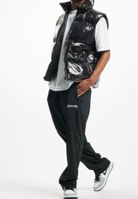 Smanicato imbottito nero lucido, maniche corte, abbinato a pantaloni neri larghi con logo bianco e sneakers nere e grigie. Tessuto liscio.