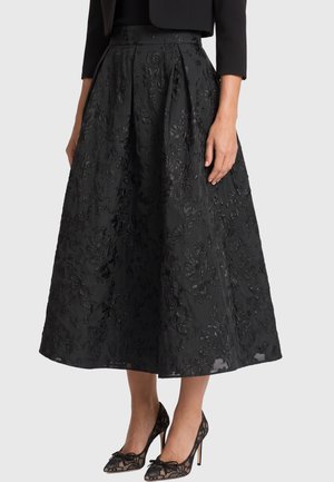 Schwarzer floraler, strukturierter Midirock, kombiniert mit schwarzen Spitzen-High Heels und einer schwarzen kurzen Jacke vor hellem Hintergrund.