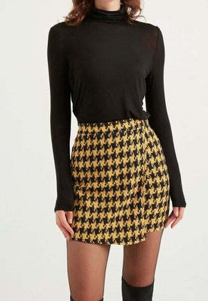 Žena nosící černý rolák s dlouhým rukávem a žluto-černou mini sukni s vazbou "houndstooth" spolu s průhlednými černými punčochami.