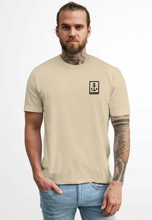 Beige T-Shirt mit kurzen Ärmeln, ausgestattet mit einem schwarzen Ankerdesign und Text im linken Brustbereich. Glattes Material, lässige Passform.