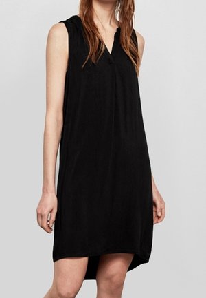 Vestido negro sin mangas hasta la rodilla con dobladillo ligeramente asimétrico, usado por una persona que está de pie contra un fondo claro y liso.