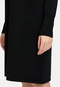Robe noire en tricot, avec des manches longues et une silhouette droite. Le tissu est lisse avec une texture subtile et un ourlet fini.