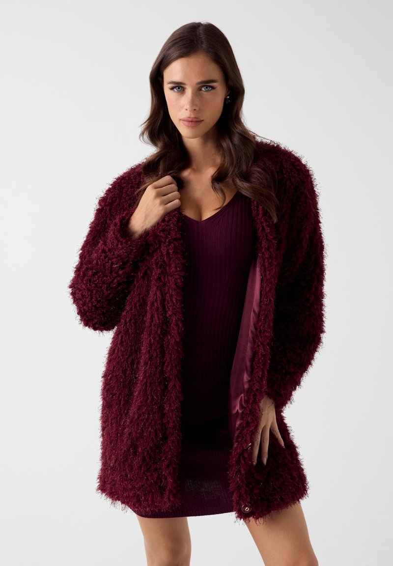 Bordeauxroter Cardigan mit flauschiger Textur und offenem Vorderteil. Aus einem weichen, gemütlichen Stoff. Wird über einem gerippten, dunkelvioletten Kleid getragen.