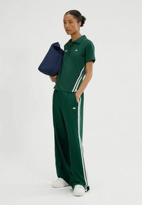 Groene polo met bijpassende trainingsbroeken met witte zijkanten. Model houdt een marineblauwe tas vast en draagt witte sneakers. Glad, atletisch materiaal.