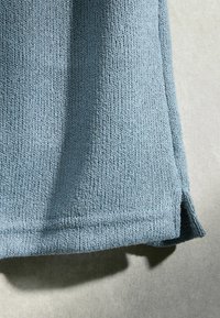 Gros plan sur un ourlet en tissu tricoté bleu clair avec une petite fente latérale sur fond clair neutre.