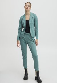 Tyrkysový blazer s límečkem a jediným knoflíkem, ve spojení s odpovídajícími tapered joggery s páskem na stažení v pase. Černý top a robustní černé boty.
