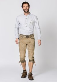 Hellgraues Langarmhemd mit einem geknöpften Kragen, kombiniert mit beigen Lederhosen, die Knöpfe an den Säumen und braune Schuhe haben.