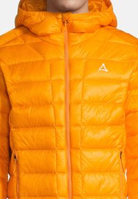 Puffjacka i stark orange, med huva, quiltat mönster och full dragkedja. Inkluderar sidofickor med dragkedja och en logotyp på bröstet.