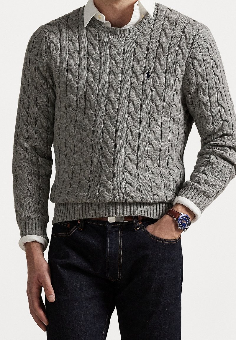 Homme portant un pull en maille torsadée gris par-dessus une chemise blanche à col, avec un jean foncé et une montre-bracelet à cadran bleu et bracelet en cuir marron.