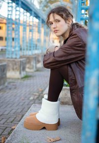 Botas de plataforma gruesas con parte superior de ante color marrón claro, puño de punto acanalado blanco y suela de goma beige, combinadas con leggings oscuros y chaqueta de cuero marrón.