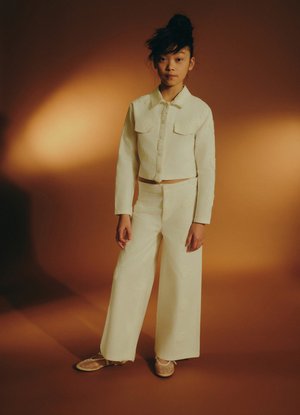 Giovane persona in piedi con espressione neutra, indossa una giacca color crema con bottoni, pantaloni a gamba larga e scarpe beige, su sfondo marrone caldo.