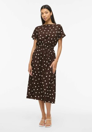 Kvinne iført en brun midikjole med hvite prikker, korte ermer, innsnevret midje, står med hendene langs siden, iført hvite sandaler.