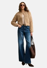 Giacca beige a trama, aderente in vita, abbinata a jeans blu a vita alta e svasati. Una borsa tote marrone completa l'outfit. Indossati occhiali da sole audaci.