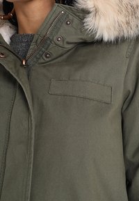 Khaki parka bunda s umělou kožešinou na kapuci, zapínání na zip a patentky. Obsahuje malou náprsní kapsu. Měkčený materiál.
