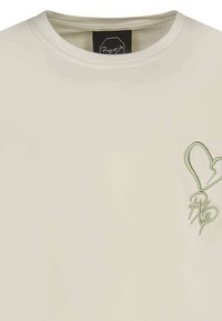 Camiseta de algodón color beige claro con cuello redondo y un diseño de corazón bordado en verde con una firma estilizada en la parte inferior.