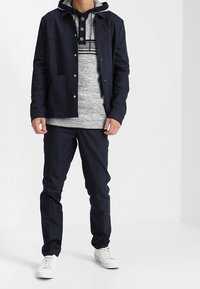 Giacca navy sopra un cappuccio grigio, pantaloni neri e sneakers bianche. La giacca ha bottoni a pressione; il cappuccio presenta un insert con motivo e bottoni.