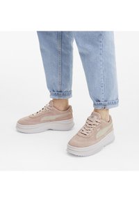 Baskets en daim rose avec une finition texturée, semelle en caoutchouc blanche et accent beige clair sur le côté, portées avec un jean bleu clair retroussé.
