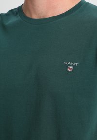 Nahaufnahme eines grünen Rundhalsshirts mit kleinem GANT-Logo und Wappenemblem auf der linken Brust.