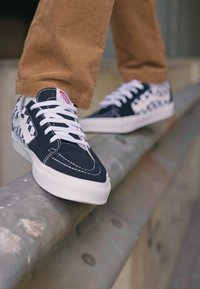 Svarte og hvite rutete sneakers med hvite snører, med overdel i semsket skinn og lerret, som står på en metallskinne, delvis skjult av bukseben.