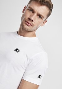 White cotton t-shirt with a crew neck, featuring a black and white logo embroidered on the left chest and matching accents on the sleeves.