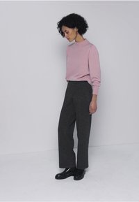 Ljusrosa långärmad tröja kombinerad med höga, gråsvarta vida byxor och svarta ankelstövlar, stående mot en neutral bakgrund.