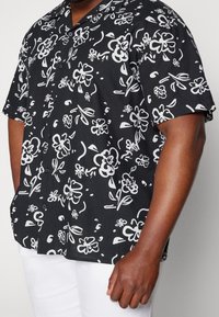 Kortsleeve svart skjorta med vita blommönster, knäppning framtill och krage. Tillverkad av lättviktsmaterial, med en avslappnad passform.