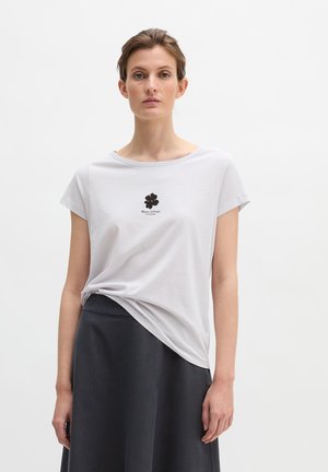 Woman wearing a white short-sleeve shirt with a black flower logo and text, paired with a black skirt, standing against a white background.