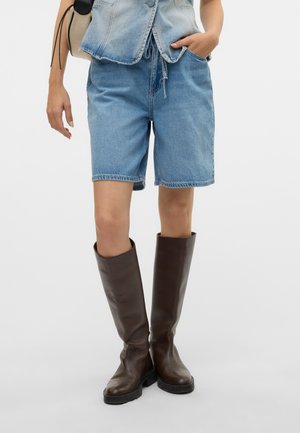 Personne portant un short en denim bleu clair, un haut sans manches en denim et des bottes en cuir marron foncé montant jusqu'au genou, debout avec une main dans la poche.