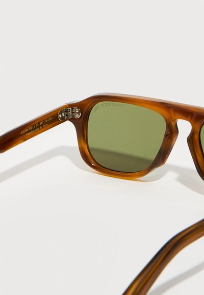 Καφέ διαφανή ορθογώνια γυαλιά ηλίου με φακούς πράσινου αποχρώματος, που δείχνουν τον μεντεσέ και την επιγραφή «Χειροποίητα στην Ιταλία» στο βραχίονα.