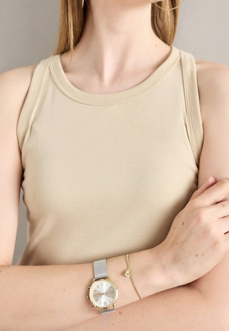 Femme portant un haut sans manches beige, les bras croisés, montre argent et or au poignet gauche, et fin bracelet en or au poignet droit.