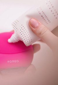 Appareil de nettoyage en silicone rose avec une surface texturée, surmonté d'une bouteille de nettoyant blanche présentant des accents à pois et un texte imprimé.