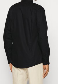 Camisa negra de manga larga con cuello abotonado, textura suave y diseño ajustado, con puños abotonados y dobladillo curvado.