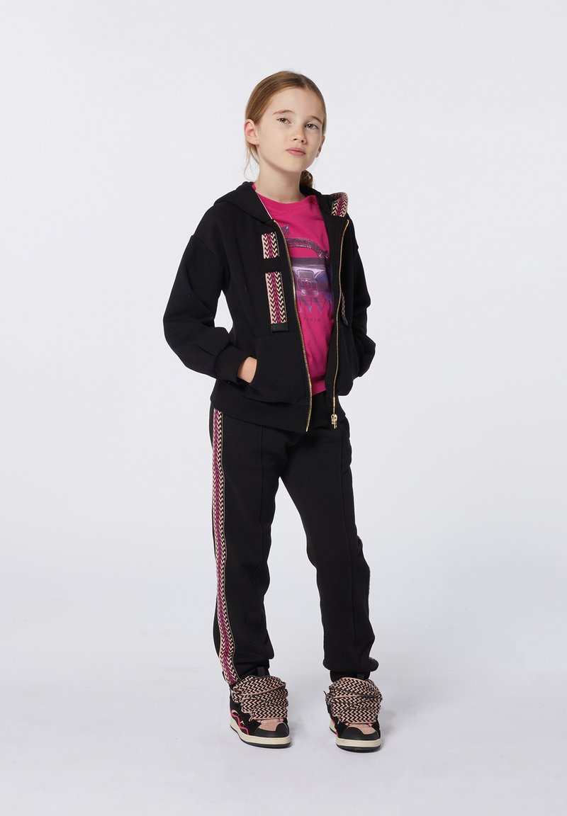 Sweat à zip noir avec des accents à motifs, associé à un pantalon noir avec une bande sur le côté. Le modèle porte un t-shirt graphique rose et des baskets à motifs.