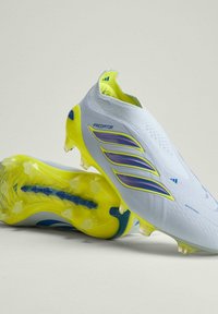 Chaussures de football Adidas Predator blanches et jaunes avec des rayures violettes et une semelle cloutée conçue pour jouer sur terrain ferme.