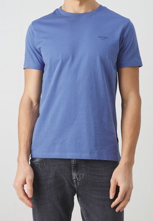 Homme portant un t-shirt bleu JOOP! Jeans à manches courtes et col rond avec un jean gris foncé, debout devant un fond clair uni.