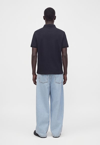 Homme debout, dos tourné, portant un polo noir à manches courtes et un jean bleu clair à jambes larges avec des chaussures noires.
