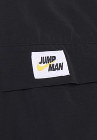 Sort stof har et hvidt, syet mærke med "JUMP MAN" i sort og en gul Nike swoosh, placeret vandret.