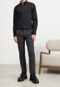 Chemise noire à manches longues avec col structuré, associée à un pantalon slim à carreaux gris et des chaussures noires à lacets avec semelles épaisses.