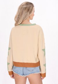 Felpa beige oversize con dettagli a forma di stella verde, maniche testurizzate e orlo a costine marrone. Tessuto morbido con un taglio rilassato.