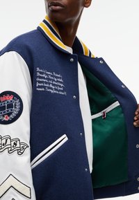 Άτομο που φοράει σκούρο μπλε πανωφόρι varsity με λευκά μανίκια, ριγωτό κίτρινο και σκούρο μπλε γιακά, κεντημένο κείμενο, μπαλώματα και πράσινη εσωτερική επένδυση.