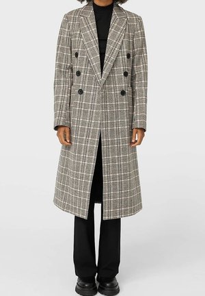 Persoon draagt een lange, dubbelgeknoopte grijze geruite jas met zwarte knopen over zwarte broek en grove zwarte schoenen.