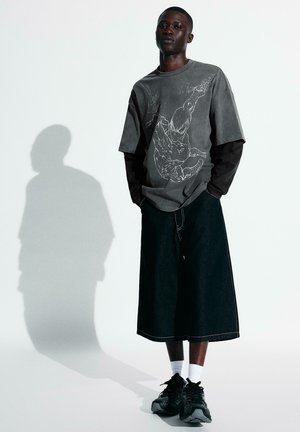 Mladý muž stojící s rukama v kapse, oblečený v nadměrné tričko s motivem Spider-Mana, široké černé šortky, bílé ponožky a černé tenisky, na pozadí bílé zdi.