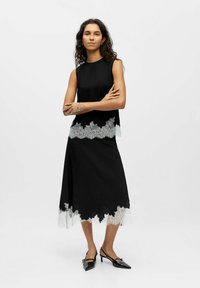 Robe noire sans manches, avec un ourlet en dentelle gris clair. Tissu lisse, longueur au genou, associée à des chaussures noires à bout pointu.